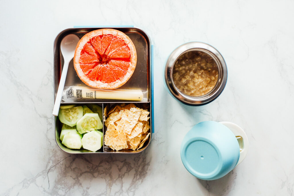 Oatmeal in a thermos with a grapefruit, cucumber slices, cheese stick, and tortilla chips on the side in a bento box.