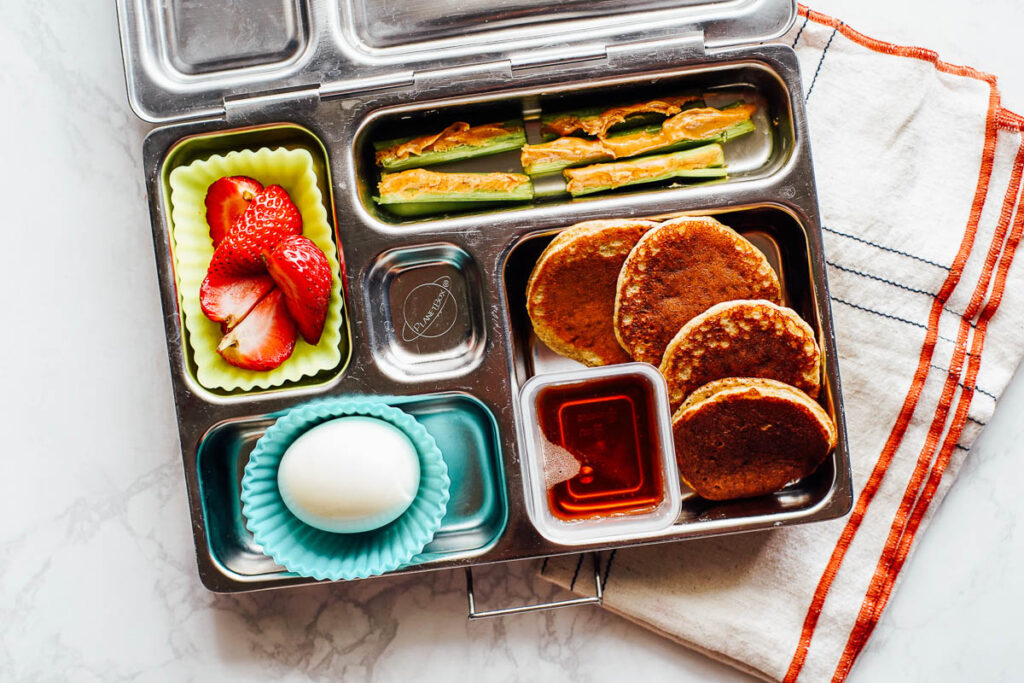 Mini pancakes with maple syrup, hardboiled egg, strawberries, and celery with peanut butter in a lunchbox.