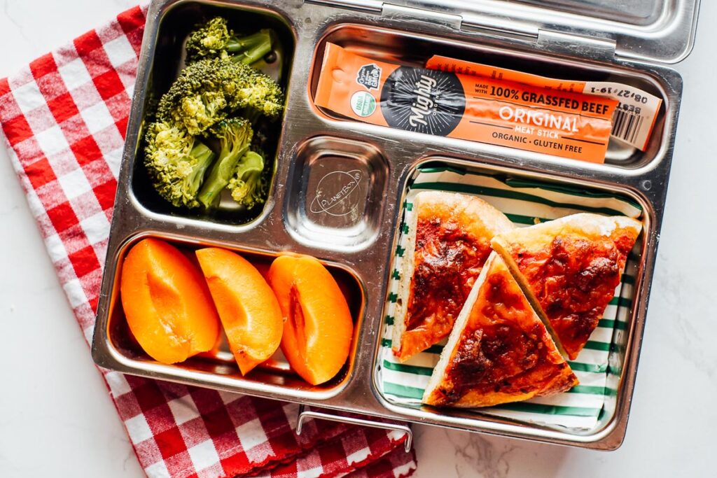 Pizza in a lunchbox with fruit, broccoli, and a meat stick.