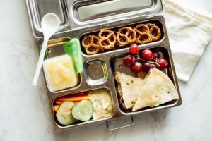 Cheese quesadilla with grapes, cucumber and carrot slices with hummus, applesauce, and pretzels in a lunchbox.