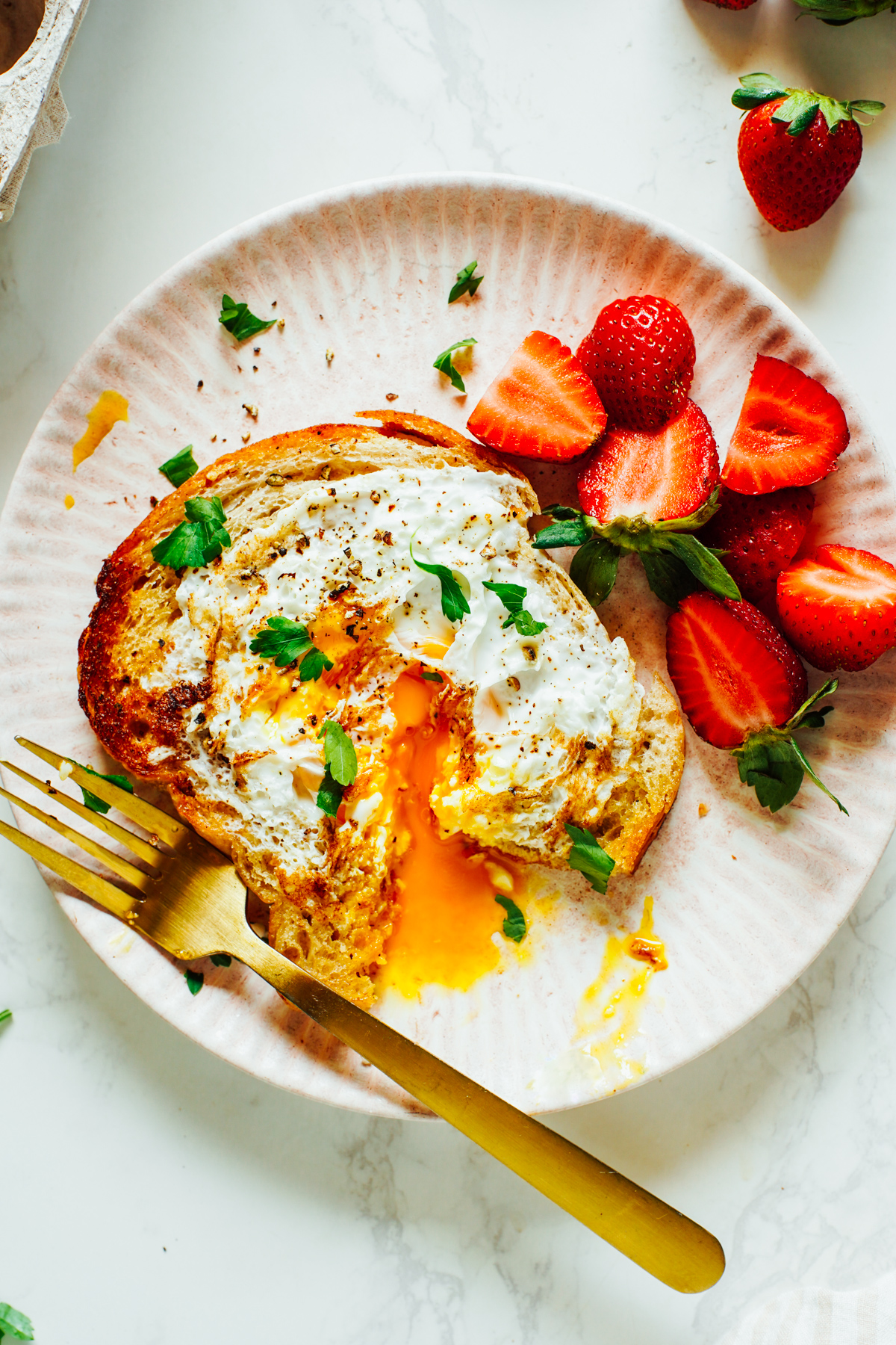 Egg cooked in toast, cracked open with the egg yolk running out on a pink plate with strawberries.