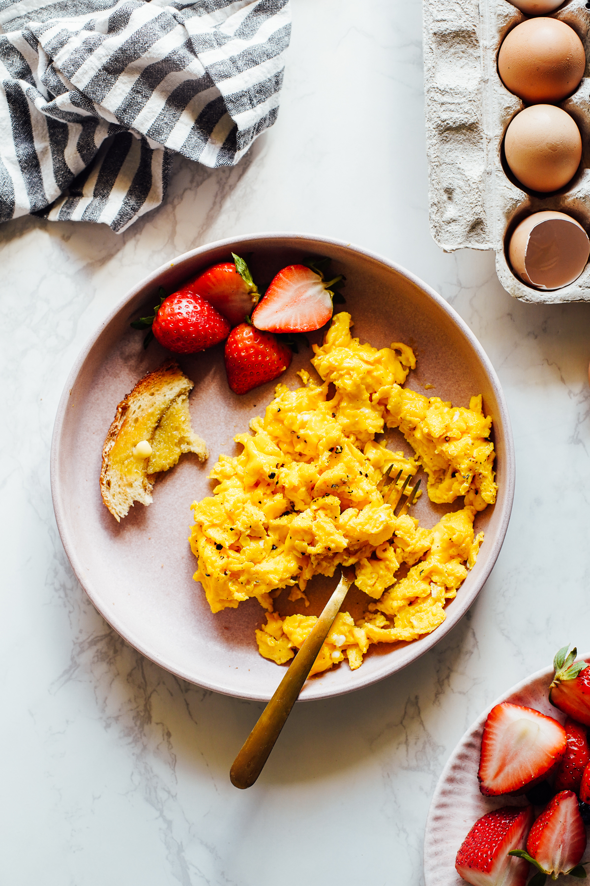 Scrambled eggs on a pink plate with strawberries and cracked eggs in a egg carton on the side.