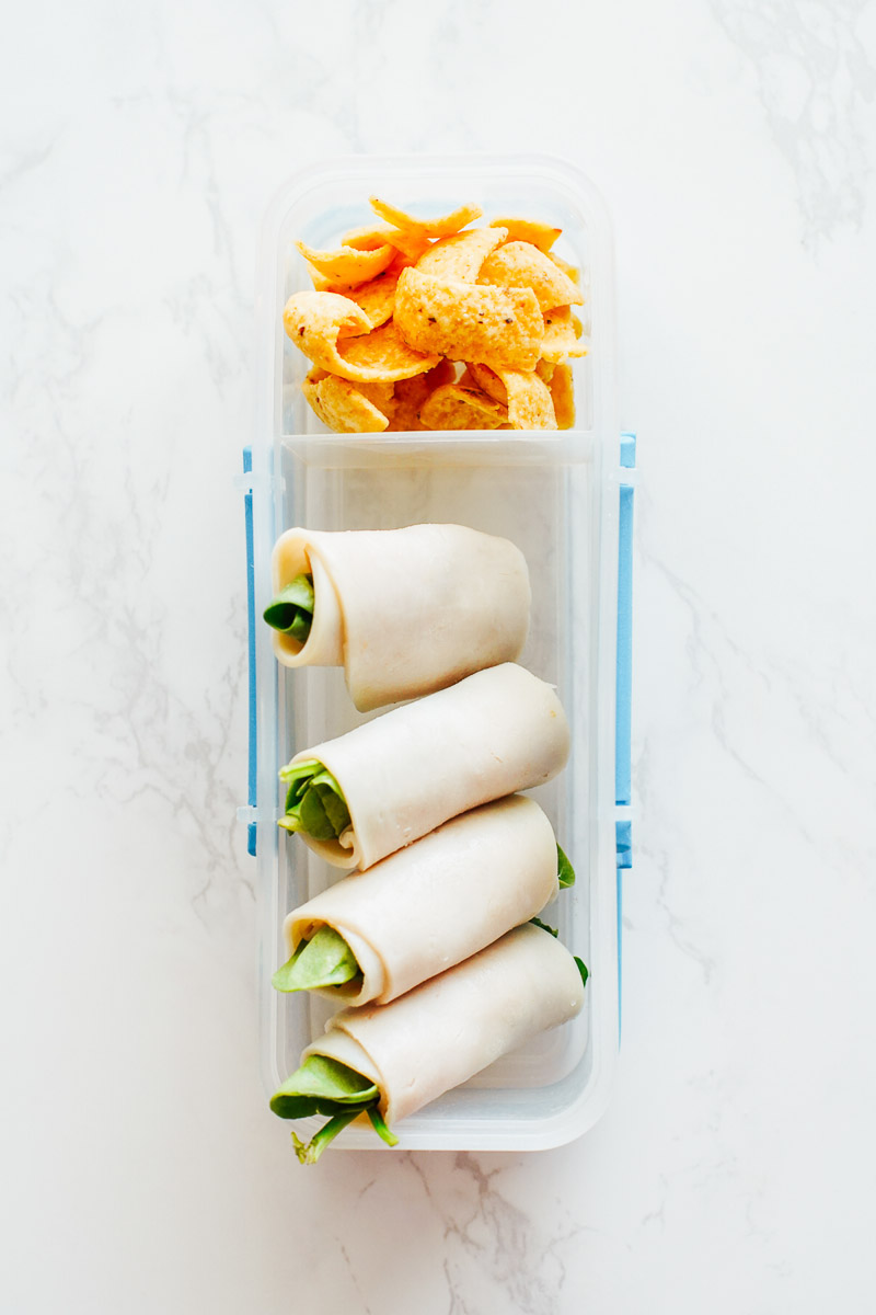 Turkey slices rolled with spinach and corn chips in a plastic snack container.