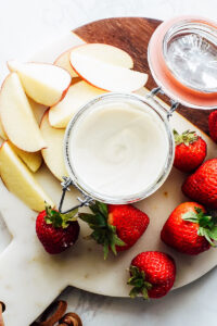 Yogurt dip for fruit in a glass bowl on a cutting board with strawberries and apples.